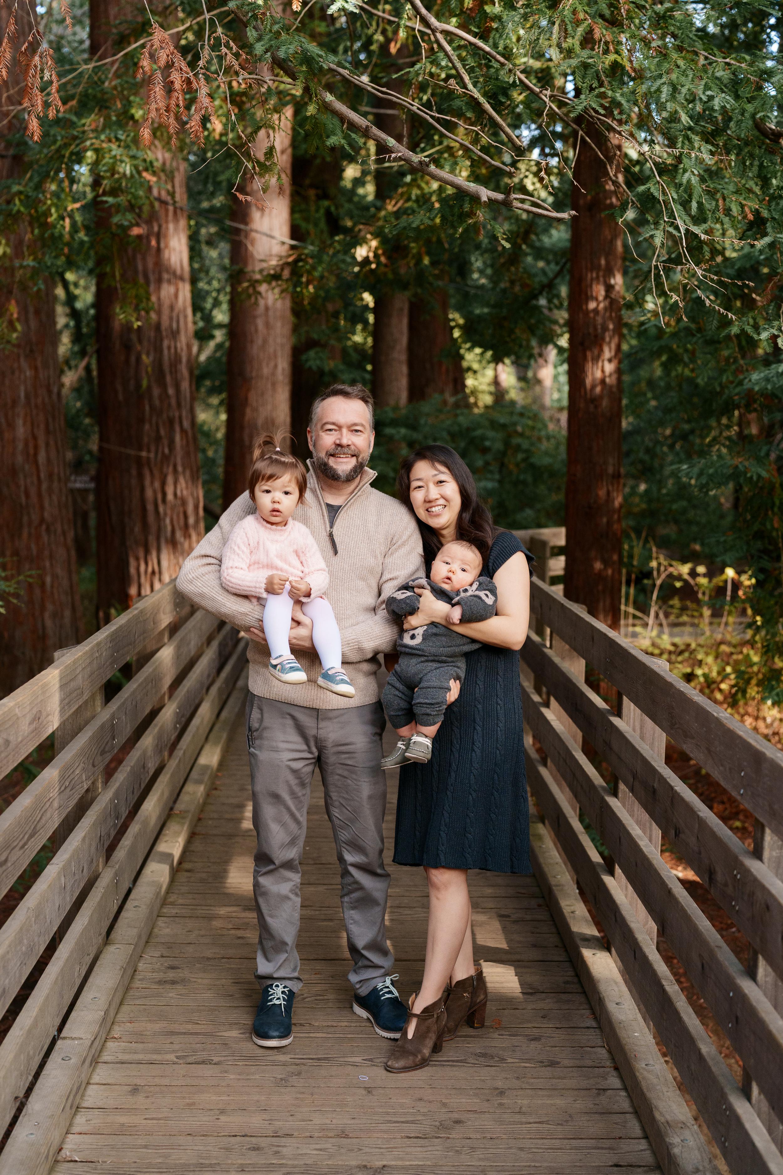 Bay Area family portrait of parents holding toddler daughter in pink and baby boy in gray on a wooden bridge among redwood