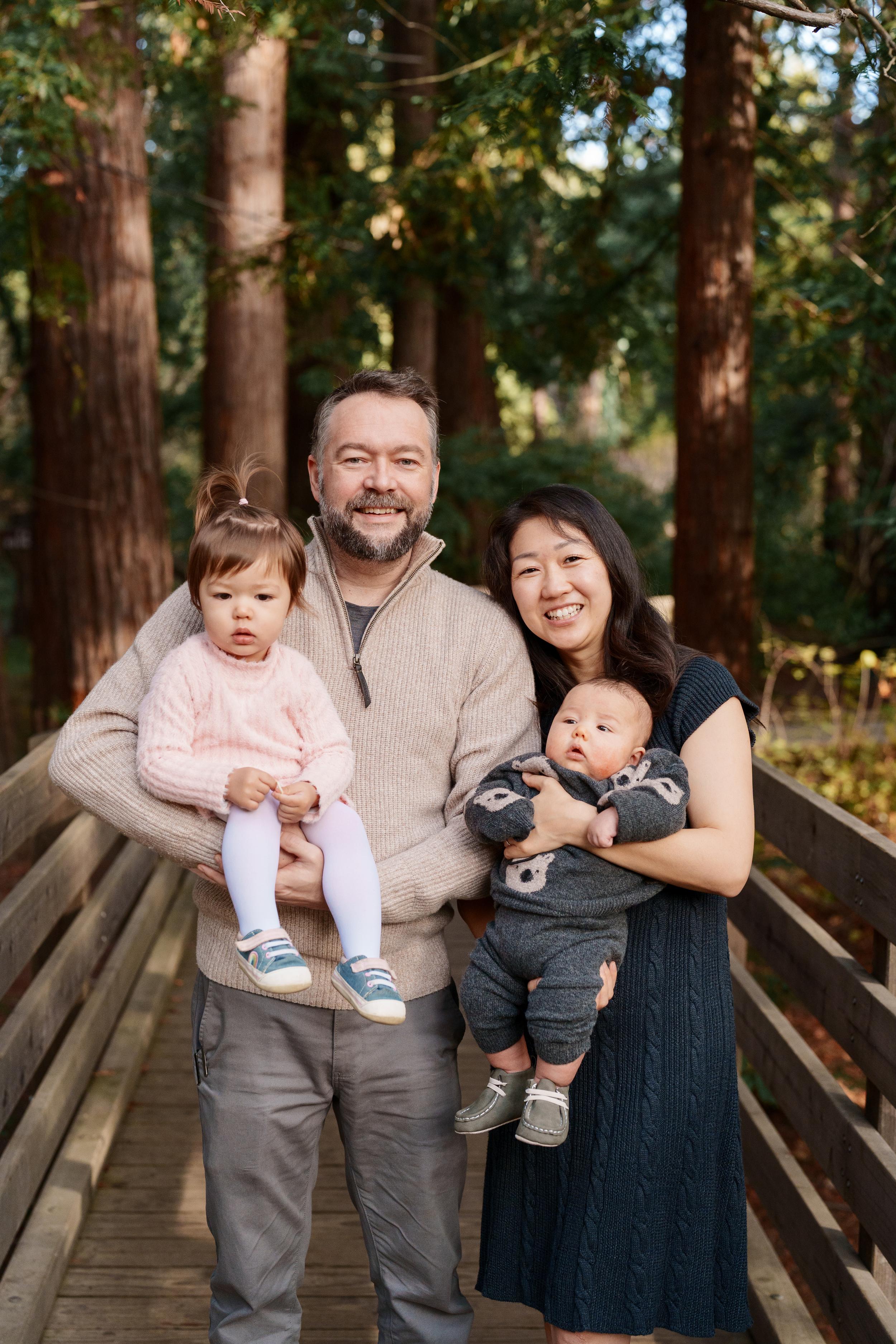 Bay Area family portrait of parents holding toddler daughter in pink and baby son on a wooden bridge among redwoods.