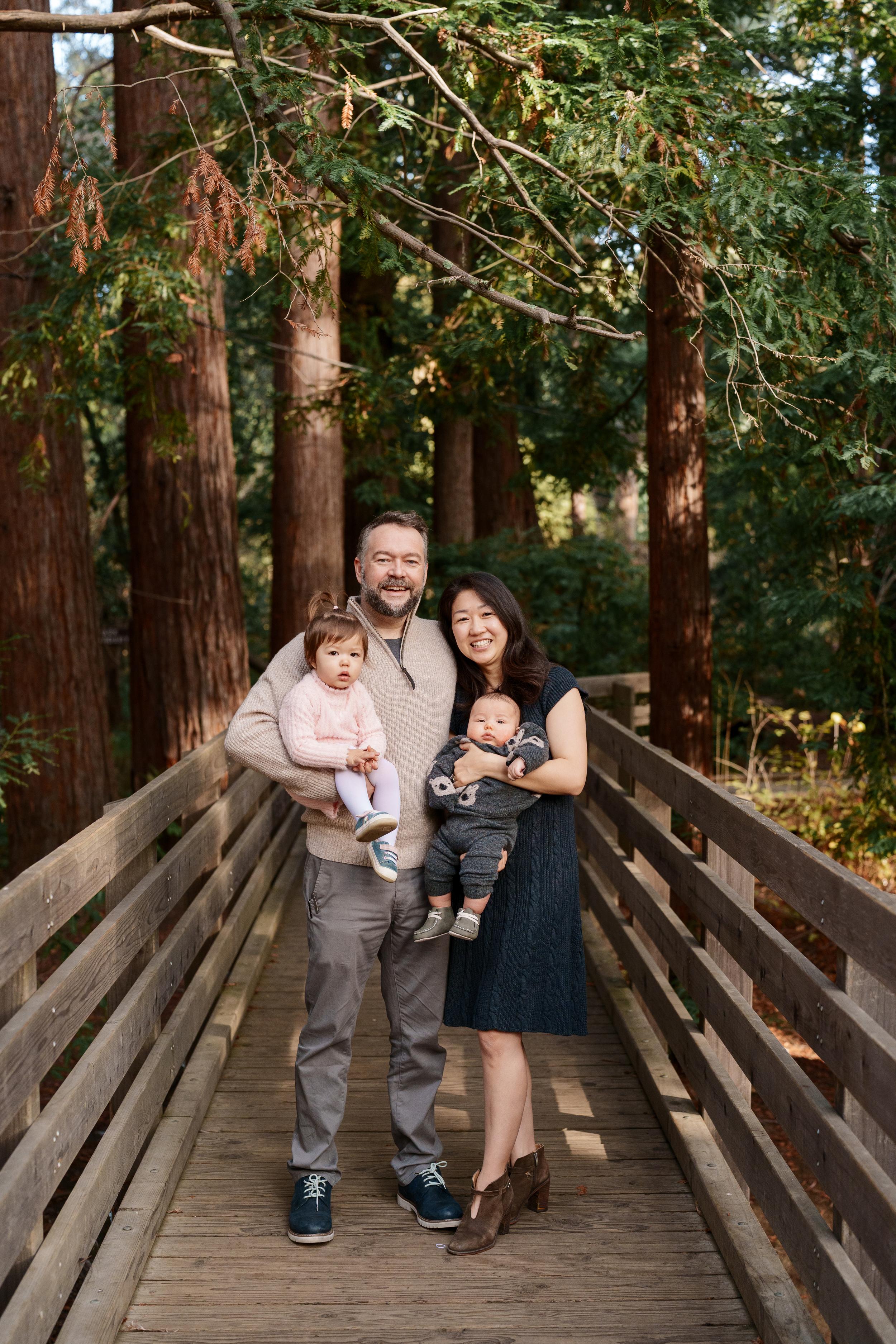Bay Area family portrait of parents holding toddler daughter and baby son on a wooden footbridge among towering redwood