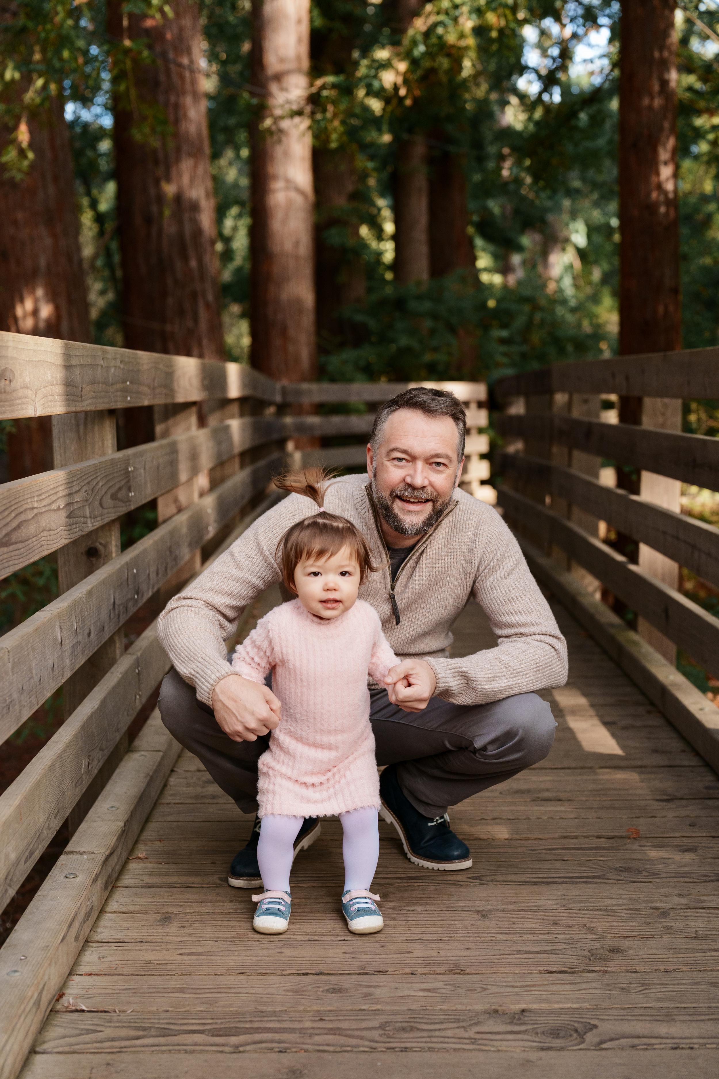 Bay Area family portrait of a bearded dad in a tan sweater crouching beside his toddler daughter in a pink dress on a wooden