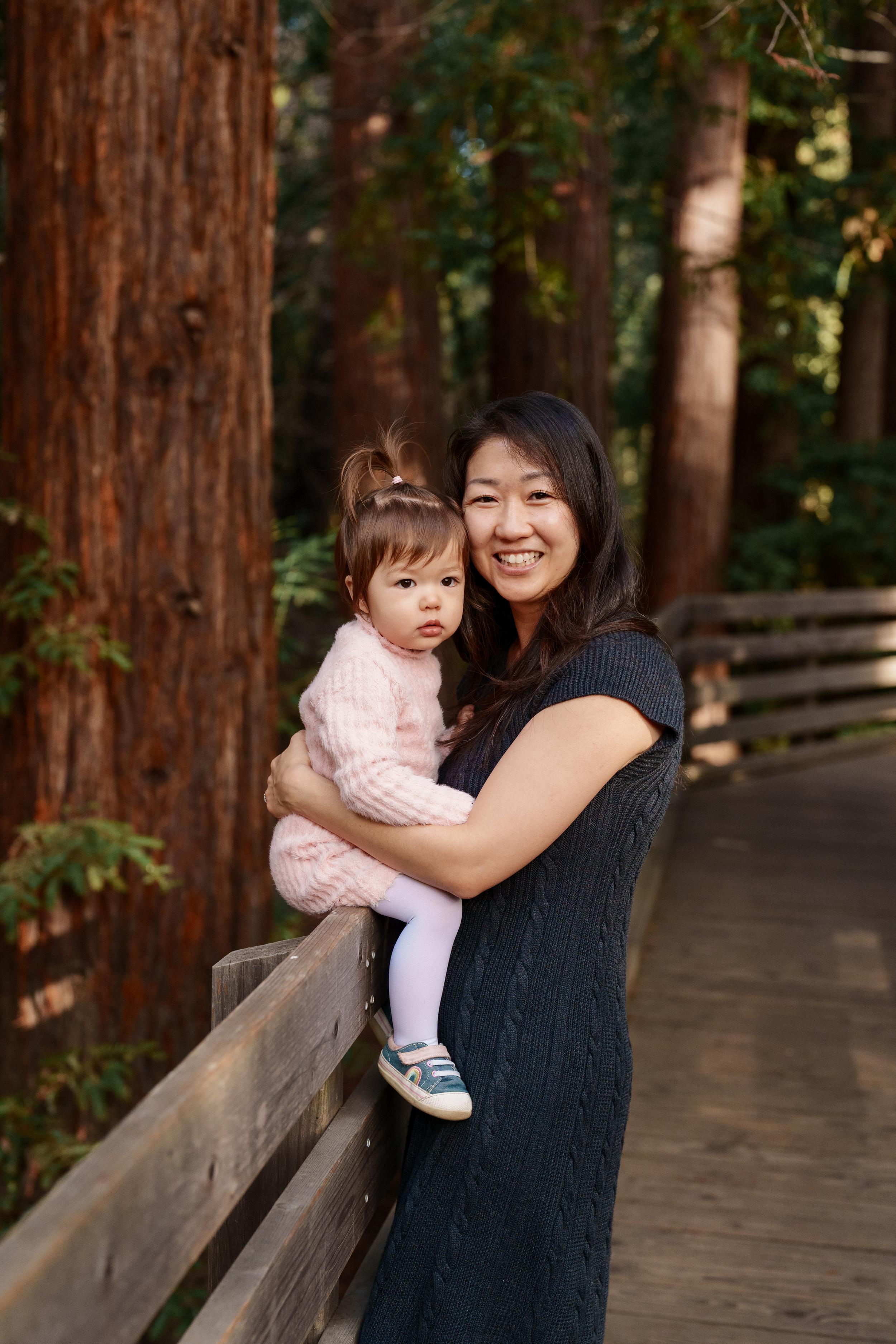 Bay Area family portrait of a smiling mother in a navy knit dress holding her toddler daughter in a pink sweater on a redwood