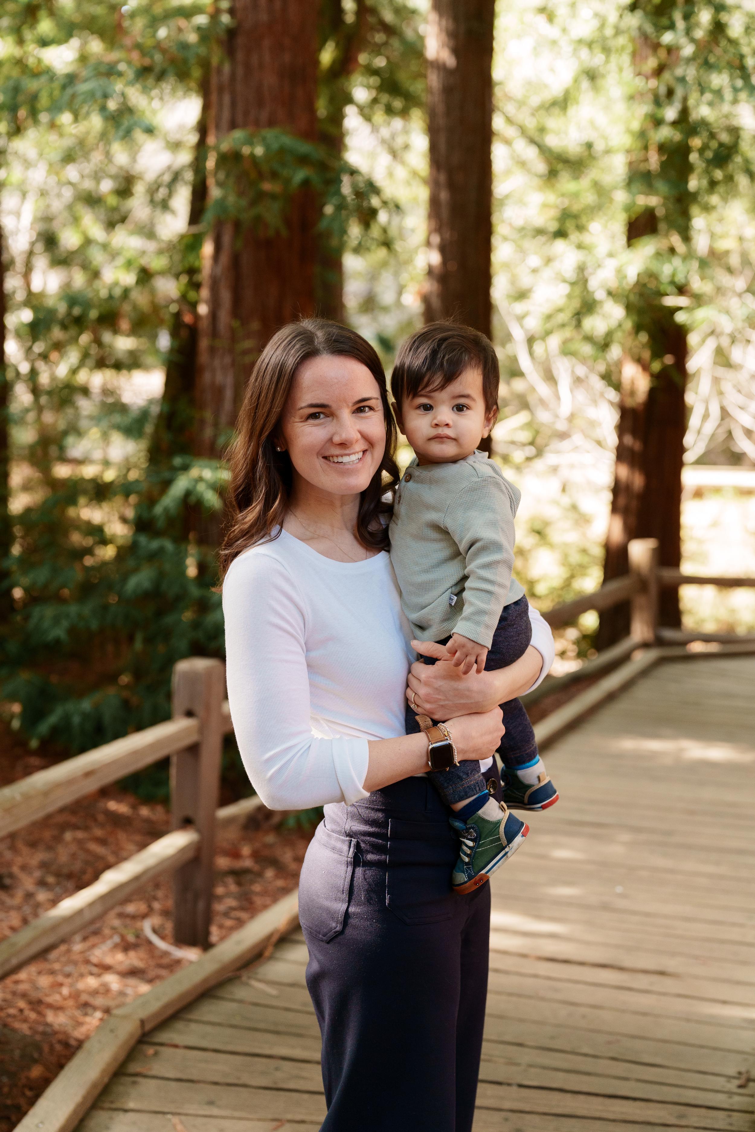 Bay Area family portrait of a smiling mom in white top holding her toddler son in sage sweater on a wooden boardwalk among