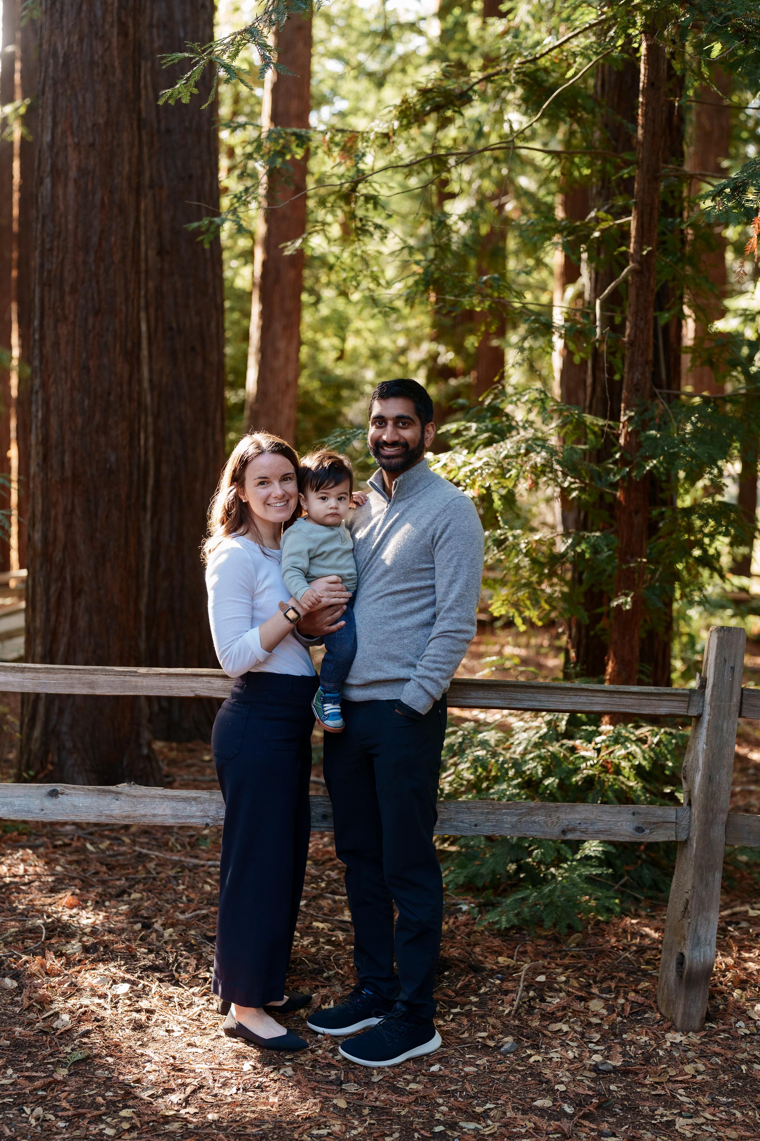 Bay Area family portrait of parents holding their toddler son by a wooden split-rail fence in a sunlit redwood grove.
