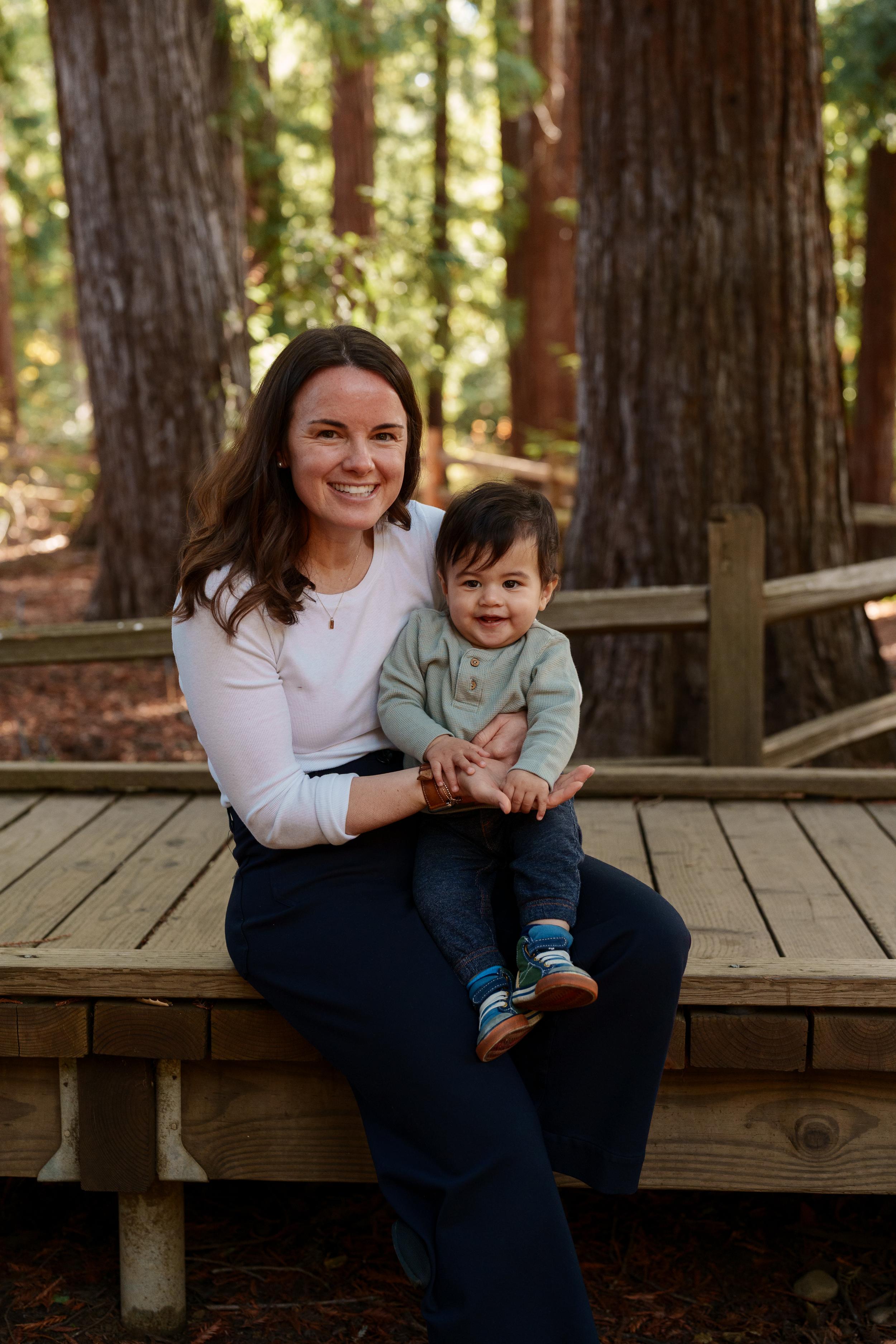 Bay Area family portrait of a smiling mother in a white top and navy pants holding her toddler son on a wooden deck among