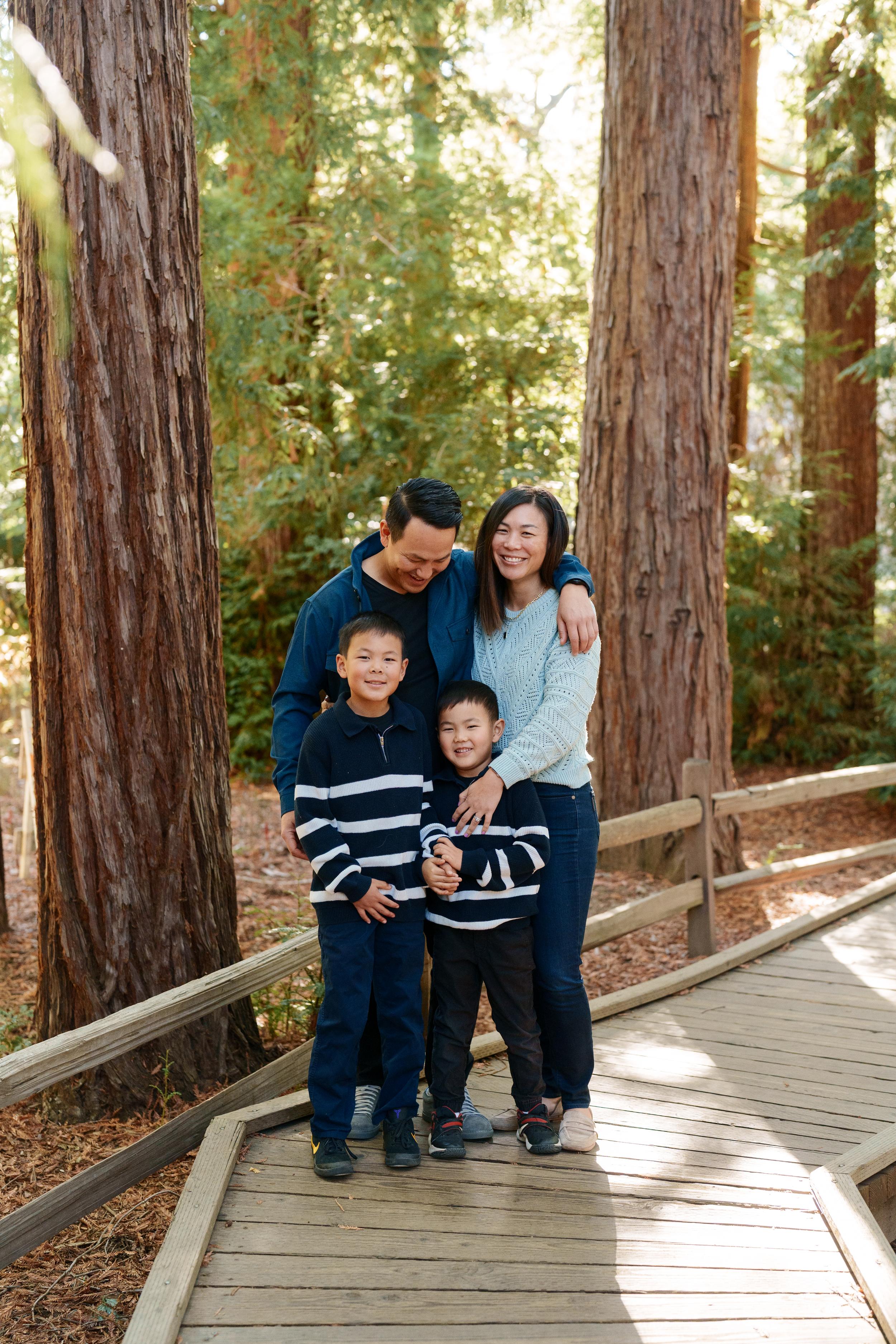 Bay Area family portrait of parents and two young sons in navy and blue sweaters hugging on a sunlit redwood grove boardwalk.