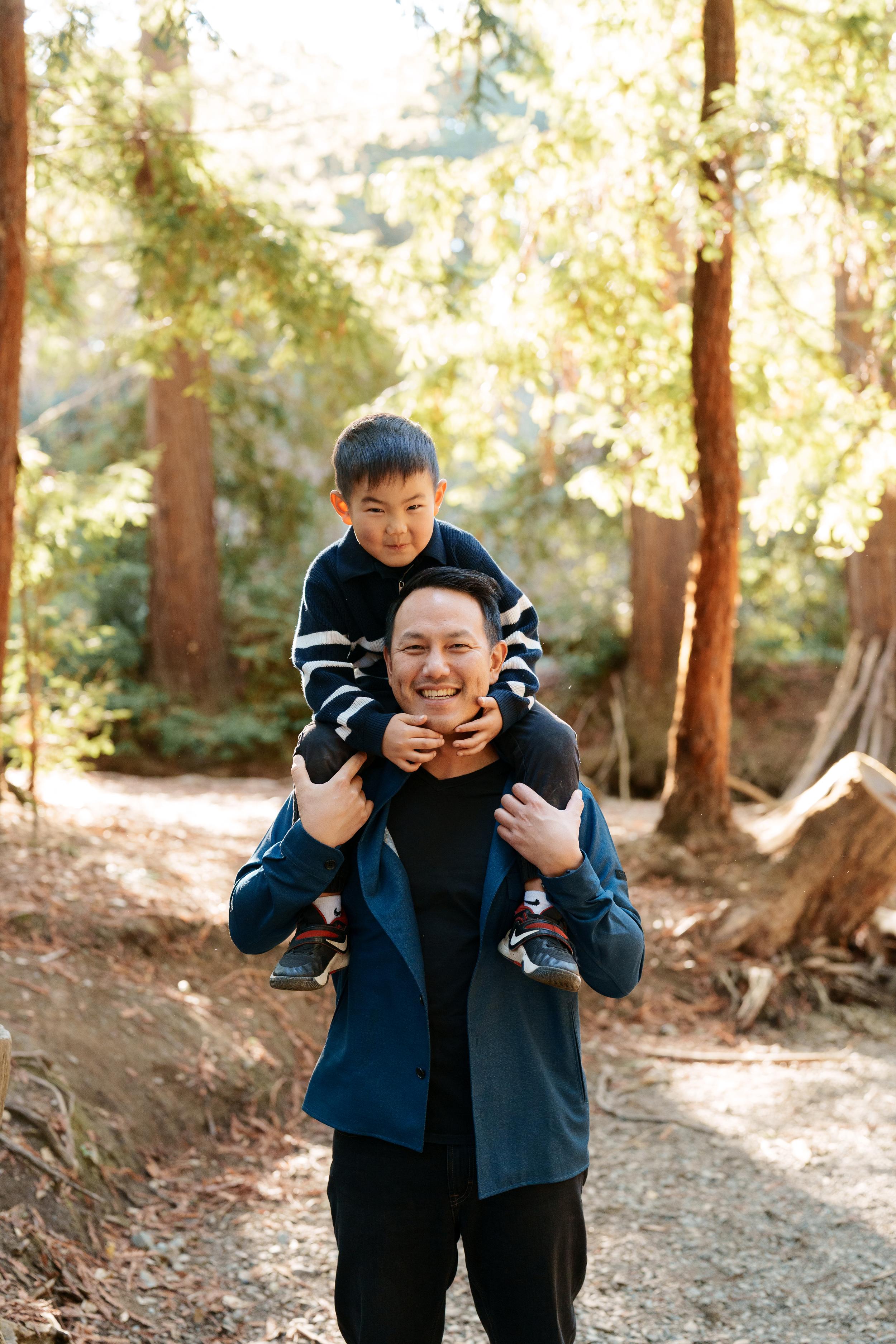 Bay Area family portrait of a smiling dad in a blue jacket carrying his young son on his shoulders along a sunlit redwood